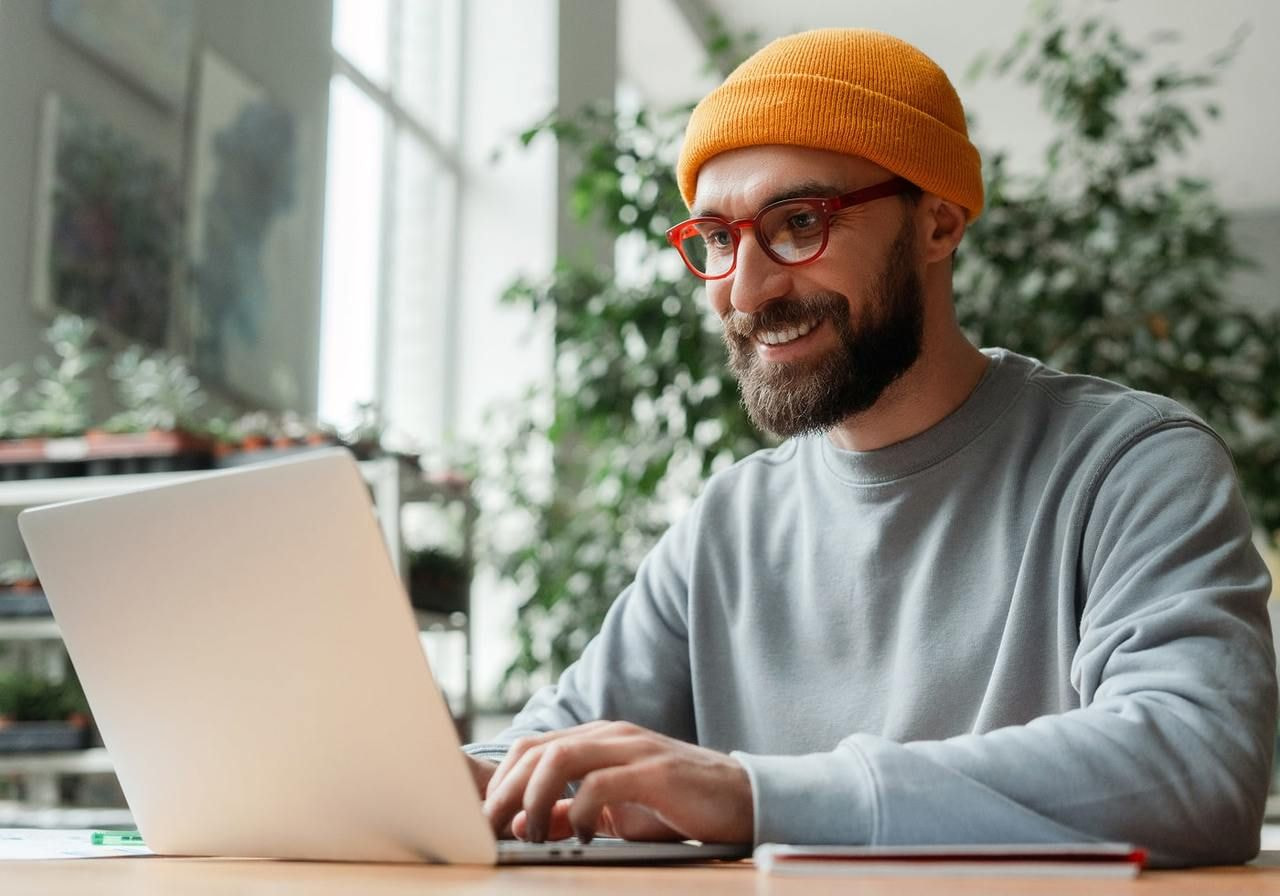 A man in an orange beanie and red glasses is smiling while using a laptop. He wears a gray sweater.